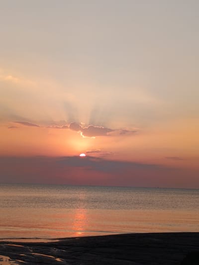 This is a photograph of a sunset over the ocean at Jangkar, Indonesia. The sun is partially obscured by clouds, casting rays of light through them. The sky is a gradient of warm colors, transitioning from a soft orange near the horizon to a pale peach and then a light beige at the top. The ocean reflects the warm hues of the sunset, creating a shimmering path of light on its surface. In the distance, a small sailboat can be seen on the horizon. The foreground shows a dark, sandy beach, possibly with some tidal pools. The overall mood of the image is peaceful and serene, evoking the feeling of a tranquil evening by the sea. The time of day is clearly sunset. There is no visible text in the image.