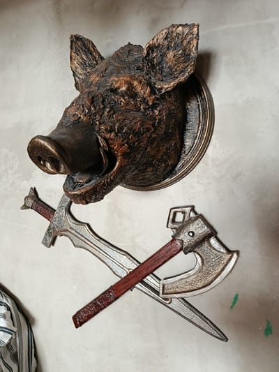 This media file features a mounted trophy of a boar's head, rendered in a dark, textured material with bronze-like highlights. The boar's mouth is open, revealing its tusks. Below the boar's head, a sword and an axe are displayed, crossed over each other. The sword has an ornate hilt and a patterned blade. The axe has a wooden handle wrapped in what appears to be leather, and a metallic head with intricate detailing. The background is a neutral-colored, textured wall. A portion of a striped garment is visible in the lower left corner. The overall impression is one of a decorative display, possibly in a home or a themed establishment. There is no visible text in the image. The setting appears to be indoors, with the lighting suggesting it could be daytime. No specific emotional cues are evident beyond the fierce expression of the boar trophy. The location context provided is Smarhon, Belarus.