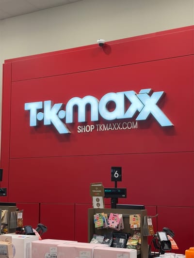This media file captures an interior view of a retail store, identified as T.K. Maxx, in what appears to be a checkout or service area. The dominant feature is a large, vibrant red wall that serves as a backdrop for the store's prominent light-blue, reflective logo, which reads "T.K. Maxx" in bold, capital letters. Below the main logo, smaller white text states "SHOP TKMAXX.COM".
To the right of the logo, a retail counter area is visible, equipped with a black digital display showing the number "6," likely indicating a till number or queue position. Below this, another small screen displays "Welcome." The counter is stocked with various small merchandise items, including greeting cards with visible text such as "HAPPY Birthday!" and "TREASURE." Packaged goods are also displayed, with visible text snippets like "Down," "SA MOSS," and "G." Several cardboard boxes, some pink, are stacked on the right side of the frame, with "COLLA" visible on one. A card payment terminal or scanner is also present on the counter.
A small white dome security camera is mounted on the red wall in the upper left portion of the image. The ceiling, visible in the upper left, is light-colored. The overall setting suggests a commercial environment in Luton, United Kingdom. No people are visible, and the time of day is indeterminate due to artificial lighting.