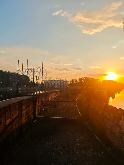 This is a medium shot of a sunset over a body of water in Vodnik SNT, Russia. The sun is a bright orb on the right side of the frame, casting a warm orange glow across the scene. The sky is a gradient of blue and orange, with scattered clouds. In the foreground, there is a weathered stone wall with barbed wire fencing along the top. A metal gate is visible in the wall. Behind the wall, trees line the water's edge. In the distance, across the water, a large building with arched windows can be seen. The water reflects the vibrant colors of the sunset. There are no people or animals visible in the image.