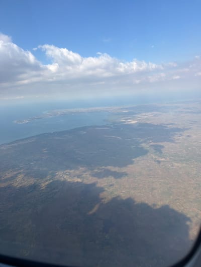 The media file is an aerial photograph taken from an airplane window, looking down at a landscape. The scene is divided horizontally, with the upper portion dominated by a bright blue sky filled with fluffy white clouds. A hint of the moon is visible in the sky. The lower portion of the image shows a terrestrial landscape, appearing to be a mix of dry, brown terrain with patches of green vegetation. A body of water, likely a river or a bay, is visible cutting through the land. The coastline is irregular, with what appears to be a small settlement or town visible on a peninsula or island. The light suggests it is daytime, with the sun casting shadows from the clouds onto the land, indicating a sunny but somewhat hazy day. The overall impression is one of looking down on a rural or semi-developed area from a significant altitude. The image is oriented vertically, making it appear as if the airplane is descending or ascending sharply.