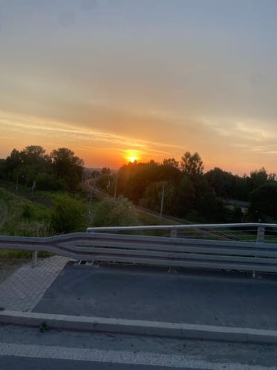 This media file depicts a sunset scene with a railway line winding through a wooded area. The sun is low on the horizon, casting a warm, orange glow across the sky and illuminating the clouds with golden hues. The trees are dense and green, suggesting a natural, possibly rural, setting. A metal railing and a paved surface, possibly a walkway or roadside, are visible in the foreground, indicating that the vantage point is from a raised structure overlooking the landscape, such as a bridge or overpass. The railway tracks are clearly visible, curving into the distance, with overhead electrical wires and posts indicating it's an active line. The overall mood is serene and peaceful, evoking the beauty of nature at the end of the day. The time of day is clearly sunset, and the weather appears clear with some cloud formations catching the light. There is no visible text in the image.