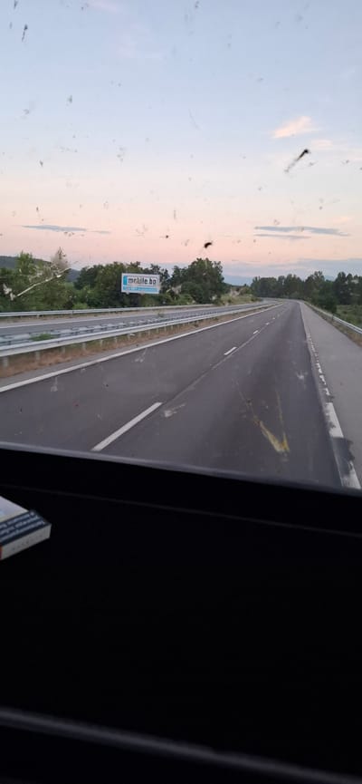 A long shot captures a highway in Blajievo, Bulgaria, under a pastel twilight sky. The road ahead curves gently to the right, bordered by metal guardrails and lush green trees. A billboard for "mobile.BG" stands prominently on the left side of the road. The foreground shows a dark vehicle interior, with a portion of a cigarette pack visible on the lower left. The image appears to be taken from a moving vehicle, with specks of dirt or debris scattered across the foreground of the shot.