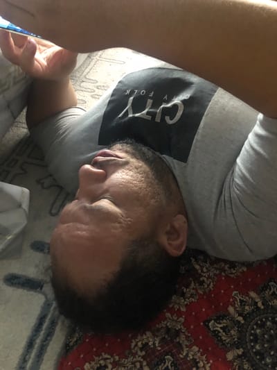 A person is lying down, holding a mobile phone in their left hand. They are wearing a grey t-shirt with a black graphic on the front that says "CITY FOLK". The person's face is visible, and they have a beard. The background appears to be a rug with a red and white pattern. No specific details about the city of Khujakent, Uzbekistan, can be determined from this image.