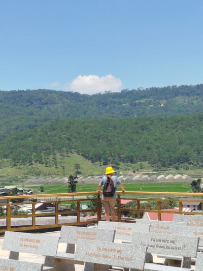 In the city of K'Rèn, Vietnam, a person wearing a yellow hat and a backpack stands at a railing, looking out over a valley. The person is dressed in a blue t-shirt and beige shorts. In the background, a lush green mountain range is visible, dotted with trees. Below the mountains, there are greenhouses and fields of crops, suggesting agricultural activity. In the foreground, there are stone benches inscribed with text, possibly memorial stones. The sky is a clear blue with a few scattered clouds. The overall scene depicts a scenic overlook with elements of nature and human development.