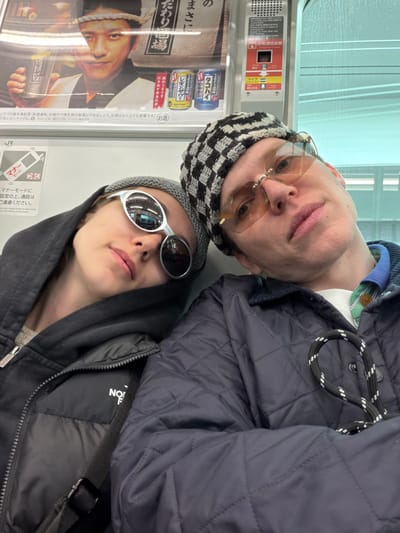 The image shows two people posing in what appears to be a train or subway car. A man is wearing a checkered beanie and tinted glasses, with a quilted jacket visible in the background. Below him, a person with a grey beanie and silver sunglasses is visible. Behind them, an advertisement features a man's face next to canned drinks and Japanese text. There is also a sign with the JR logo encouraging passengers to use manners and phones in silent mode.