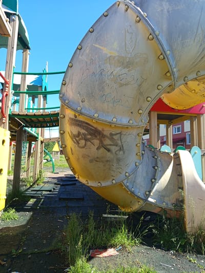 A close-up shot shows a section of a bright yellow and silver children's playground slide under a clear blue sky. The metal parts of the slide are covered in scratches and graffiti. To the left of the slide, a wooden and green metal playground structure with various levels and access points is visible. The ground beneath the playground is covered with a dark, textured surface, interspersed with black rubber mats and patches of overgrown green grass and weeds. In the background, a residential building with light-colored walls and red accents can be seen. The image appears to be taken outdoors on a sunny day.