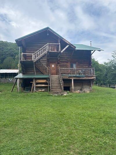 This image shows a multi-story log cabin in Azhek, Russia. The cabin is constructed from large, roughly hewn logs and features a green metal roof. Intricate wooden railings adorn the balconies and stairs, showcasing detailed craftsmanship. A prominent staircase leads up to the second-floor balcony. The exterior appears weathered, suggesting age and exposure to the elements.
The setting is rural, with a well-maintained grassy lawn in the foreground. Behind the cabin, a dense forest with lush green trees can be seen. A wooden fence is visible in the background, bordering the property. The sky is partly cloudy, with patches of blue visible among the white clouds. A thin chimney pipe extends from the roof of the cabin.
There are no people visible in the image, and no specific activity or event is taking place. The lighting suggests it is daytime, possibly late morning or afternoon, given the brightness and the contrast of light and shadow on the cabin. The overall impression is one of rustic charm and a peaceful, natural environment. No visible text is present in the image.