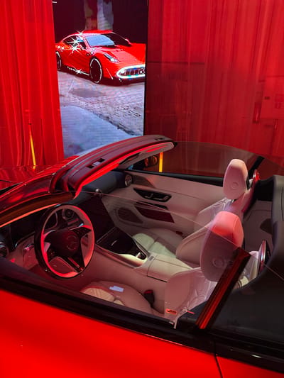 The scene is an illuminated display within a location in Herentals, Belgium. A dark-colored Mercedes-Benz convertible sports car, with its roof retracted, occupies the foreground, its exterior reflecting significant red ambient light. The interior features pristine white leather seating and trim, with protective plastic film visible on the lower sections of the seats. A multi-function steering wheel bearing the Mercedes-Benz emblem is visible on the driver's side. A transparent barrier separates the viewer from the vehicle's interior. In the background, a large vertical LED screen displays a stylized red sports car, resembling a Ferrari F12berlinetta, with prominent white outlines and sparkle effects, set against a textured ground. This screen is flanked by draped red curtains. No individuals are present in the image.