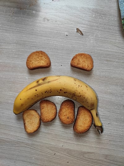 A banana and six oval-shaped crackers are arranged on a light gray wooden surface. The banana is positioned horizontally, curving upwards slightly, with its peel showing brown spots. Five crackers are placed below the banana, forming a curved line that mimics the shape of a mouth. Two additional crackers are positioned above the banana, suggesting eyes. The surface appears to be a tabletop or floor with visible wood grain and some minor scuff marks. There are no people visible in the image. This image does not contain any information that could be used to determine its location in Nefteyugansk, Russia.