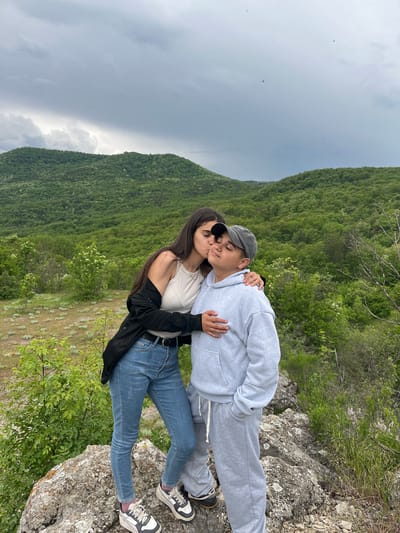 A couple is standing on a rocky outcrop with a densely forested hillside in the background. The sky is overcast with dark grey clouds. The woman on the left has long dark hair, is wearing a light-colored tank top, blue jeans, and a black jacket slung over her shoulder. She has sunglasses on her head and is embracing and kissing the other person on the cheek. The person on the right is wearing a grey hoodie, grey sweatpants, a grey baseball cap, and athletic shoes. They are standing on a rocky surface covered with moss and some low-lying vegetation. The picture was likely taken outdoors, potentially on a hiking trail or scenic viewpoint. The location is Kamchia, Bulgaria.
