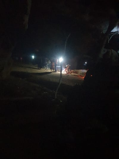 At night, a group of figures gathers around a brightly lit area. Some appear to be working, possibly using tools that emit sparks. The scene is dimly lit with several bright light sources. The surroundings are dark, obscuring details beyond the immediate illuminated space. A branch is visible in the foreground. The atmosphere suggests a work site or an emergency situation.