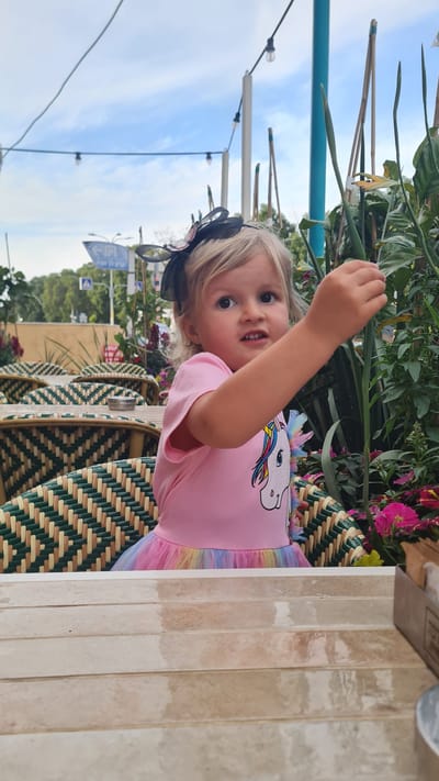 A young child wearing a pink unicorn shirt and a headband stands near an outdoor table. The table features woven chairs. The setting appears to be an outdoor cafe or restaurant, with plants and flowers visible in the background. Street signs indicate the location as Kiryat Gat, Israel. The sky is partly cloudy.