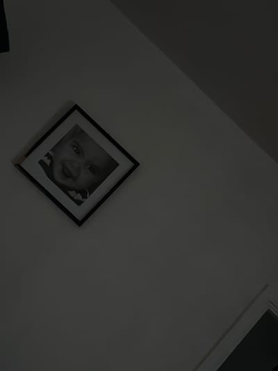 A black and white framed photograph of a baby is hanging on a wall in a dimly lit room. The photograph is square and tilted so it appears like a diamond. The baby in the photograph is smiling and looking directly at the camera. The background of the photograph is dark. The wall the photograph is hanging on is white. The image is taken from a low angle, looking up at the photograph. There is a shadow cast on the wall to the right of the photograph. The room appears to be a bedroom or living room.