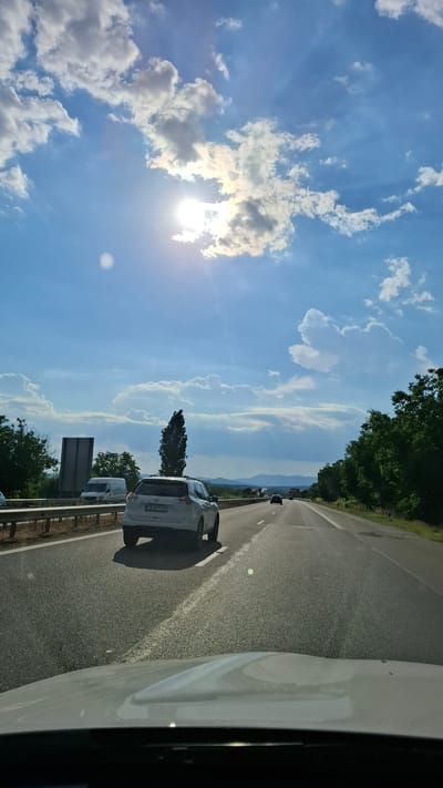 This media file depicts a scene from a vehicle traveling on a highway, likely in Kalugerovo, Bulgaria. The perspective is from inside a white vehicle, with its side visible on the right. The highway stretches ahead, with several other vehicles visible, including a white SUV in the foreground and a white van further down the road. The road is paved with white lane markings.
The sky is a prominent feature, a brilliant blue with scattered white cumulus clouds. Sunlight streams through the clouds, creating bright rays and a strong glare. The time of day appears to be mid-morning or early afternoon, given the bright sunlight and clear sky. The weather seems to be warm and sunny, ideal for travel. Lush green trees line the right side of the highway, with a hint of rolling hills or mountains in the distant background, suggesting a rural or semi-rural setting.
The activity taking place is travel on a highway. The overall mood is serene and somewhat expansive, likely due to the open sky and the feeling of movement. There is no visible text in the image that can be read clearly, except for a license plate on the white SUV in the foreground which appears to read "DB 2611 CE".