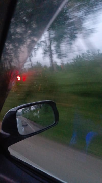 The image shows a view from inside a moving vehicle. The perspective is through the driver's side window and includes the side-view mirror. The side-view mirror reflects a road and trees, suggesting the vehicle is on a road in a rural or semi-rural area. In the foreground, out of focus, are parts of the car interior, including a dark-colored dashboard and possibly a door panel. To the left of the side-view mirror, and also out of focus, is a red digital display, possibly from another vehicle, with the numbers "153" visible. The exterior view is blurred due to motion, but it appears to be a cloudy or overcast day with green foliage and trees. There is no specific information in the image that points to the city of Зуй, Russia.