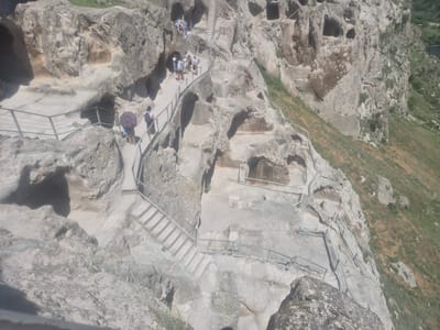 This media file depicts a scenic view of the Vardzia cave monastery in Georgia. The scene is dominated by a steep, rocky cliff face carved with numerous caves, which once served as monastic dwellings and churches. A series of stone staircases and pathways, equipped with metal railings, wind their way up the cliff, providing access to the various levels of the ancient complex.

Several people are visible navigating these paths, suggesting a tour or a visit by tourists. They are dressed in casual, warm-weather clothing, and one individual is holding an umbrella, hinting at a sunny, possibly warm, day. The overall atmosphere appears peaceful and exploratory, with visitors taking in the historical and natural beauty of the site.

The natural landscape surrounding the monastery is also evident, with patches of dry grass and green vegetation clinging to the slopes, indicating a natural environment. In the distance, a body of water, possibly a river, can be seen, adding to the scenic backdrop. The lighting suggests it is daytime, with sunlight illuminating the rock formations and pathways. The image captures the grandeur of the ancient architecture seamlessly integrated into the natural geological formations. No visible text is present in the image.