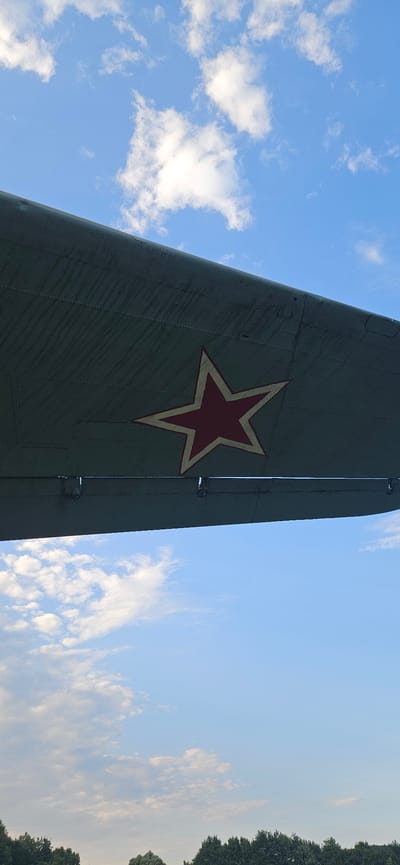The image shows a portion of an airplane wing with a red star insignia, set against a blue sky with scattered white clouds. The wing is painted a muted green and exhibits signs of wear, with visible panel lines and rivets. A dark line, possibly a tree line, is visible at the very bottom of the frame. The location is identified as Vyšný Komárnik, Slovakia, which is known for the Dukla Pass, a significant historical site related to World War II.