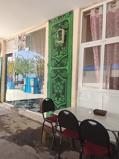 The image captures a storefront in Sariosiyo, Uzbekistan, featuring a glass-fronted establishment with a green decorative mat hanging on the exterior wall. Inside, stacks of blue and yellow boxes are visible through the glass, along with trees and reflections of the surroundings. In front of the store, a white marble-topped table is set with a small basket and a shaker of salt, accompanied by two chairs with black frames and red upholstery. The ground outside is a cracked concrete surface, with a large puddle of water reflecting the sky and surrounding structures. A portion of a window with pink curtains is also visible on the right side of the image.