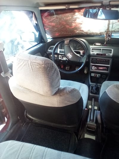 The image shows the interior of a car, likely taken from the passenger seat, looking towards the driver's side. The focus is on the car's dashboard, steering wheel, gear shift, and front seats. The seats are covered in a light-colored fabric with a textured pattern, and the lower portions are black. The dashboard is dark gray, with air vents, a radio or CD player, and instrument cluster visible. The steering wheel is black and features a Honda emblem in the center. A rearview mirror hangs from the ceiling. The scene appears to be set during the daytime, given the light coming through the windshield, which is partially obscured by a reflective sunshade. The overall impression is of a well-used but functional car interior. There is no visible text in the image. The context provided indicates the location is Grigoriopol, Moldova.