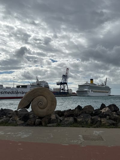 The image presents a daytime scene at the port of Puerto del Rosario, Fuerteventura, Spain, under an overcast sky. In the immediate foreground, a paved promenade, featuring reddish-brown asphalt on the left and grey concrete with an embedded metal grate on the right, extends across the frame. Directly beyond the promenade, a line of dark, irregular rocks forms a boundary. Centrally positioned on these rocks is a large, light-toned sculpture of a spiral shell, oriented horizontally. The midground features the calm, grey-blue water of the harbor. On the left, a large white ferry, partially displaying "osmediterrán" in red lettering on its hull, is visible, possibly docked. Adjacent to the ferry, a prominent dark blue port crane with a red apex stands, surrounded by stacked shipping containers on the dock. To the right, a large white cruise ship, identifiable by its yellow funnel, is either docked or at anchor. The background consists entirely of a dense, grey cloud cover, with minimal patches of brighter sky. No individuals are present in the scene.