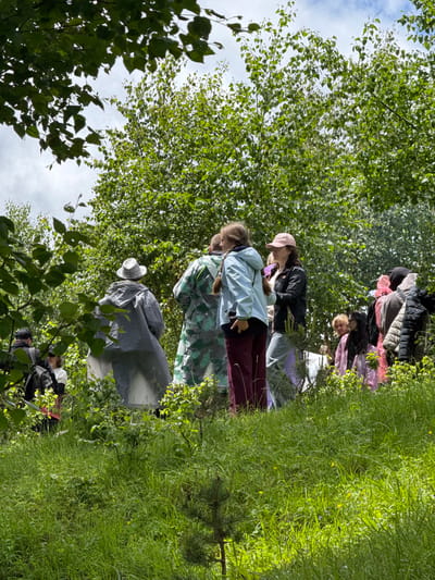 In an outdoor scene amidst lush green foliage and trees, a group of people are gathered on a grassy incline. The background is dominated by dense green trees and a cloudy sky. In the foreground, tall grass with some small yellow flowers covers the ground. Several individuals are visible, facing away from the camera or in profile, suggesting they are engaged in an activity or conversation. One person in the center wears a gray hooded raincoat and a patterned hat. To their right, another person is seen in a green camouflage-patterned garment. Other individuals are dressed in casual outdoor clothing, including jackets, hoodies, and trousers. The overall impression is that of a gathering in a natural, possibly forested, environment in the city of Nizhnyaya Teberda, Russia.