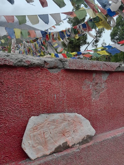The image shows a close-up view of a weathered, textured red wall in Jispa, India. A rough, light-colored stone with carved inscriptions is placed on the ledge in front of the wall. In the background, strings of colorful prayer flags are visible against a cloudy sky and distant mountains, suggesting a spiritual or cultural setting. The wall itself shows signs of wear and tear, with chipped paint and exposed concrete. The carving on the stone appears to be in a script common to Tibetan Buddhism, possibly Om Mani Padme Hum.