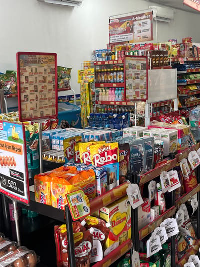 The image is a medium shot of a brightly lit convenience store aisle, likely in Meteseh, Indonesia, given the Indonesian language on product labels and signage. The aisle is filled with various consumer goods displayed on red and white shelving units. In the foreground, to the left, a price display board shows "Rp 80.000" and "Rp 21.500" with images of eggs. Further to the left is a red display board with "PALING MURAH SEJAGAT" and product promotions.

Dominating the middle ground are stacks of packaged instant noodles, notably "Mie Sedaap" in orange packaging and "Kuaci Rebo" in yellow and red packaging. There are also boxes of milk, like "Promina" and "Buvita," and various bagged snacks. On the right side, more shelves are laden with bottled drinks, packaged foods, and household items such as cleaning supplies. Small promotional tags and flyers are attached to some products. In the background, a blue refrigerated display case is visible, and part of an air conditioning unit is mounted high on the wall. The overall environment is typical of a retail setting, with a focus on merchandise presentation for sale.