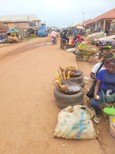 This image depicts a vibrant outdoor market scene, likely in Tangu, Nigeria. The primary subjects are people engaged in commerce and daily life. A woman is seated, holding an egg, appearing to be a vendor or customer. She is surrounded by various goods, including grilled corn displayed on makeshift grills made from tires. Nearby, a cart is laden with fruits, possibly limes or lemons.

The setting is a bustling street market, characterized by stalls and vendors selling their wares. Motorbikes and a blue truck are visible, indicating transportation and the movement of goods and people. The ground is a dusty, reddish-brown earth, typical of many Nigerian environments. The weather appears to be overcast or hazy, with a bright sky suggesting it's daytime.

The overall atmosphere is one of activity and commerce, with a focus on food preparation and sale. The grilling of corn suggests a food stall in operation. The presence of various vehicles and people suggests a busy marketplace, likely during peak hours.

There are a few pieces of visible text: "PALANG OIL LTD" is clearly printed on a building facade. Another sign reads "PALANG BARKA" above a shop. The lettering on the burlap sack is partially visible and difficult to decipher completely but appears to have blue stripes and some printed text. The general scene conveys the lively and dynamic nature of a local Nigerian market.