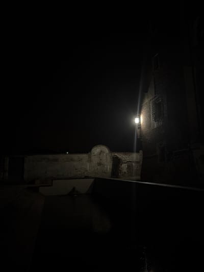 The image depicts a dimly lit, exterior scene at night, likely in the historical center of Lagnasco, Italy. The foreground is dominated by a dark body of water, possibly a canal or fountain, with small points of light reflecting on its surface.
To the left, a weathered, aged wall runs horizontally across the frame. This wall features architectural elements like a partially visible arched entryway and sections where the brickwork is exposed. The wall appears to be part of an older structure, possibly a courtyard or a protected area.
To the right, a brick building stands tall, its facade illuminated by a single, vintage-style lantern mounted on the wall. This lantern casts a bright, focused beam of light that creates a distinct streak of illumination against the dark night sky. The building itself has a window with a barred grate, suggesting historical security measures or architectural design. The brickwork appears rough and aged, consistent with older construction.
The overall atmosphere is one of quiet solitude and historical ambiance, enhanced by the nighttime lighting and the aged structures. The scene suggests a place steeped in history, where the passage of time is evident in the materials and architecture. There are no people visible in the image.