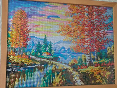 The image displays a diamond painting of a serene autumn landscape. The painting is framed in a simple wooden frame. The scene depicts a body of water in the foreground, with reflections of trees and a small house. In the background, there are lush green trees, mountains, and a colorful sky with a sunset. The painting is detailed, with each diamond representing a small point of color, creating a mosaic-like effect. The overall impression is one of natural beauty and tranquility. There is no indication of the city of Serebriansk, Kazakhstan.