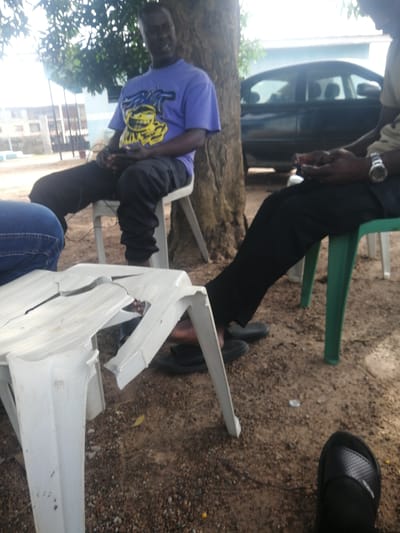 The image depicts an outdoor scene in Ajasse Ipo, Nigeria, likely in a somewhat informal setting. Three individuals are present, though their full figures are not entirely visible. One person, a man wearing a purple t-shirt with a yellow graphic and black pants, is seated on a white plastic chair, leaning against a tree. He appears to be holding a device, possibly a phone, in his hands. Another individual, also seated on a white chair, is partially visible on the left, wearing blue jeans. The third person is seated on a green plastic chair to the right, wearing dark pants and a watch on their left wrist. In the foreground, a white plastic table, visibly cracked and broken, rests on the sandy, gravelly ground. Behind the individuals, a black car is parked, and in the background, there are hints of buildings and trees, suggesting a residential or semi-urban environment. The lighting suggests it is daytime, with dappled sunlight filtering through the trees.