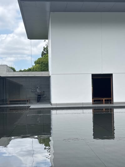 The image depicts a modern architectural complex at the 21st Century Museum of Contemporary Art in Kanazawa, Japan, viewed across a large, dark-tiled water feature that occupies the foreground.
The building on the right features a stark, multi-paneled white facade with clean horizontal lines. A large, dark, rectangular opening, possibly an entrance or deep recess, is present in the lower right section of this facade, revealing a dark interior with visible light brown wooden elements at its base.
To the left of the white facade, a dark-tinted glass wall extends across the mid-ground. This glass wall has a visible grid structure and reflects the interior, where at least two human figures are discernible. One figure, wearing a light-colored top and a face mask, is centrally reflected. Faint outlines of what appear to be light brown benches are also reflected inside. Beyond the glass wall, another section of the building with a flat, light grey roof is visible, with green trees peaking over it and in the distance.
A dark grey, flat roof overhang extends from the top left of the building against a partly cloudy blue sky. A slender, silver-colored metal rain chain hangs vertically from the edge of this overhang, disappearing towards the water feature.
A thin, grey metal handrailing runs horizontally along the edge between the building's base and the water feature. The water itself is dark and reflective, mirroring the sky with patches of white clouds and the building's structures, including the white facade, the dark opening, and the rain chain which creates a distinct wavy vertical reflection. The bottom of the water feature reveals a grid of large, dark grey square tiles. The water's surface exhibits ripples, distorting the reflections.