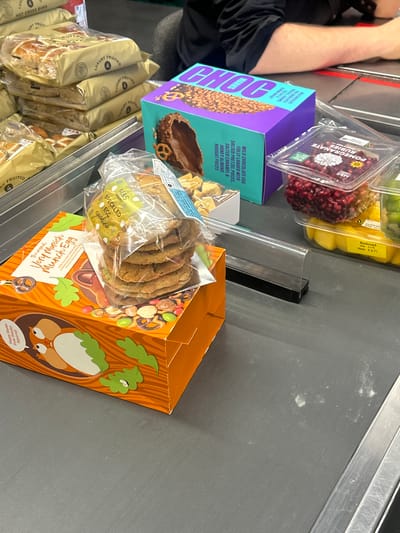 The image depicts a selection of grocery items on a checkout conveyor belt, likely in a supermarket located in Chippenham, United Kingdom, based on the branding of the products.
Visible items include:
- "LUXURY FRUITED HOT CROSS BUNS"
- "MARKS CHOC"
- "M&S SPECKLED EGG COOKIES"
- "Very Moreish Munch Egg"
The items are arranged close together and are partially obscuring each other, suggesting a recent placement on the conveyor belt in preparation for scanning at checkout.
The conveyor belt is grey, and a portion of a person's arm is visible.