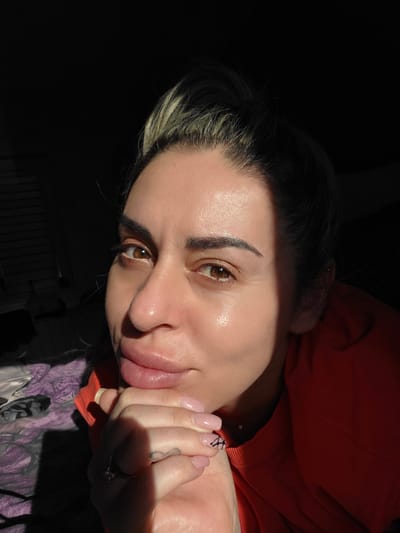 The image shows a close-up of a woman's face, with harsh shadows obscuring much of the background. The woman has her hands clasped together in front of her chin. She has brown eyes, full lips, and dark hair pulled back, with some lighter colored strands near the front. She is wearing an orange top. The lighting suggests the sun is low, casting strong highlights and deep shadows across her face. The environment appears to be indoors, possibly a bedroom, with a glimpse of a patterned blanket or comforter on the left. There is no indication of the city of Carpi, Italy.