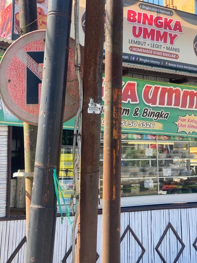 The image shows a street scene in Sungai Paring, Indonesia, featuring a food stall with prominent signage for "BINGKA UMMY." The stall appears to be selling various food items, displayed in a glass-fronted refrigerator. Several metal poles are visible in the foreground, one of which has a "no parking" sign attached to it. The overall environment suggests an outdoor commercial area with a slightly weathered aesthetic, characterized by rust on the poles and cracked paint on the sign. The signage indicates the business specializes in "BINGKA UMMY" which is described as "LEMBUT LEGIT MANIS" and "HOMEMADE KHAS BANJAR." Social media handles and a phone number are also present on the signage. In the background, other businesses and what appear to be residential structures can be seen.