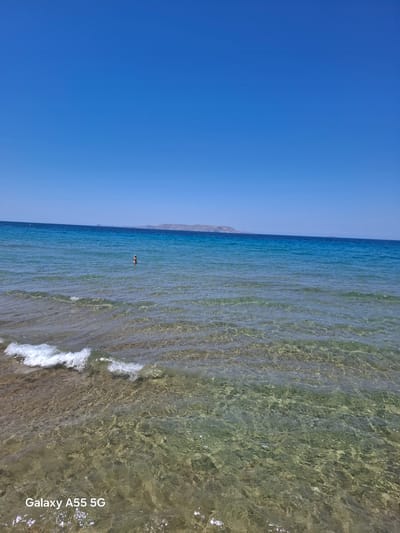 A daytime coastal scene is depicted under a clear, cloudless blue sky, occupying the upper two-fifths of the frame. The horizon line is straight, separating the sky from the sea. The sea's coloration ranges from transparent light blue in the foreground, revealing a textured sandy and rocky seabed, to a deeper turquoise, and then a dark blue in the distance. Small, white-capped waves are breaking near the bottom left, indicating a shoreline close to the viewer. A single individual is visible standing in the water, approximately waist-deep, positioned slightly left of the central vertical axis. The individual appears to be wearing dark swimwear and is facing away from the camera, or slightly to the right, with their head and shoulders above the water. In the distant background, a low, elongated landmass, consistent with the silhouette of Dia Island, is visible on the horizon, extending across a significant portion of the mid-distance. The location is a beach within the Gouves Municipal Unit, Crete, Greece, with a clear view of Dia Island. A "Galaxy A55 5G" watermark is present in white text in the bottom-left corner of the image.