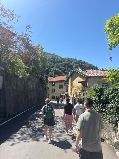 In Gittana, Italy, on a sunny day, a group of people are walking down a paved path lined with trees. The scene is dominated by a clear blue sky and lush green foliage. To the left, a hillside covered in dense trees rises sharply, with houses nestled among the greenery. Some of the houses have terracotta tiled roofs. To the right, the path is shaded by the dappled sunlight filtering through the leaves of trees.

The people are casually dressed, suggesting a leisurely activity. One person is wearing a green backpack and dark shorts. Another is in a dark t-shirt and pink and white striped shorts. Further up the path, other individuals are visible. The overall atmosphere appears relaxed and pleasant, indicative of a warm, clear day. The sun is high in the sky, casting long shadows, suggesting it is likely midday or early afternoon. There is no visible text in the image.