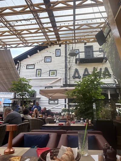 The image shows an outdoor seating area of a restaurant with a view of a white wooden building with black trim. The building has many small windows and large, decorative lettering that appears to spell "ДАНА" (Dana). A sign below the lettering reads "КРАСОТЫ" (Beauty). The roof of the seating area is made of a translucent material supported by a wooden lattice structure, with string lights hanging across it.

Several tables are set with red napkins and glassware. People are seated at some of the tables. In the foreground, a copper lamp and a basket of bread are visible on a table. To the right, a glass with a green straw sits on a table. In the background, a tree with green leaves stands in front of the building. The overall scene suggests a well-appointed outdoor dining environment in Zhukovka, Russia.