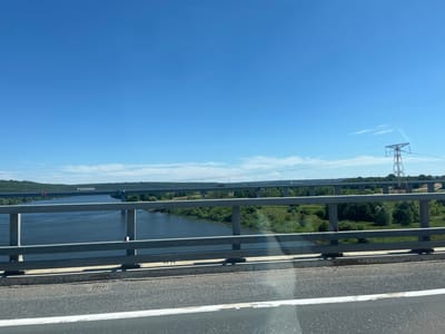 This media file is a landscape photo taken from a bridge, likely on a sunny day with a clear blue sky. The foreground features a gray metal railing and a section of asphalt road with a white line. Beyond the railing, a wide, dark blue river flows, bordered by lush green trees and vegetation on both banks. In the distance, multiple bridges span the river. The closest bridge in the mid-ground has several vehicles, including a white truck, visible on its deck. Further in the background, another bridge is visible, with a single white truck on it. A tall, lattice-style power line tower stands to the right of the image, with power lines stretching across the horizon. The overall scene suggests a rural or semi-rural location with significant infrastructure, possibly in Zasur'e, Russia. There is no visible text in the image. The time of day appears to be daylight, given the bright sky and strong shadows. The weather seems to be fair and warm.