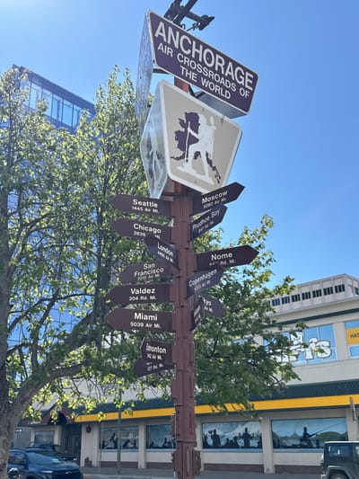 The image shows a signpost in Anchorage, Alaska, United States, indicating various cities and their distances. The main sign reads "Anchorage Air Crossroads of the World." Attached to the post are several arrow-shaped signs pointing in different directions with the names of the cities and their distances, including Seattle (1445 Air Mi.), Moscow (5282 Air Mi.), Prudhoe Bay (847 Rd. Mi.), Chicago (2839 Mi.), London (4490 Air Mi.), San Francisco (2015 Air Mi.), Valdez (304 Rd. Mi.), Miami (5039 Air Mi.), Nome (5 Rd. Mi.), Copenhagen (4313 Air Mi.), Edmonton (1442 Air Mi., 127 Rd. Mi.) and Tokyo. The signpost is surrounded by trees with green leaves, and behind it are buildings. A light blue sky is visible at the top of the frame. A building with murals is in the background to the right. A vehicle is visible to the right.