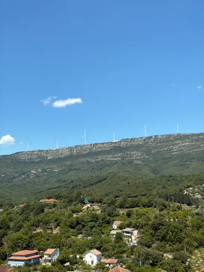 The image depicts a scenic, mountainous landscape under a clear blue sky with scattered clouds. The foreground features a dense, green forest covering rolling hills. Nestled within the foliage are several residential buildings, some with prominent red tile roofs, and others with lighter colored exteriors. A winding road is visible in the lower left, leading to a cluster of houses. Further up the mountainside, more sparsely vegetated slopes are dotted with what appear to be smaller structures or terraces. At the crest of the highest hill, a line of wind turbines stands tall against the horizon, indicating a wind farm. The overall impression is one of rural tranquility and a harmonious blend of nature and renewable energy infrastructure.