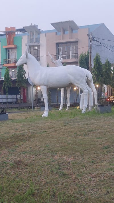 In the city of Pusakarakyat, Indonesia, a prominent white sculpture of antelopes stands in the foreground on a grassy area. The sculpture depicts several antelopes standing together, their forms rendered in a smooth, matte white finish. The main antelope in the foreground is facing left, with its head held high and a single horn visible. Behind it, other antelopes are partially visible, creating a sense of depth.

In the background, a modern building complex rises with a distinctive architectural style. The building features a combination of colors, including green, orange, and beige, with large windows and balconies. Trees line the area in front of the buildings, providing a touch of nature to the urban landscape. Electrical wires and a lamppost are also visible, indicating the presence of infrastructure. The sky is overcast, suggesting a cloudy day. The overall scene captures a public space with artistic elements and contemporary architecture.