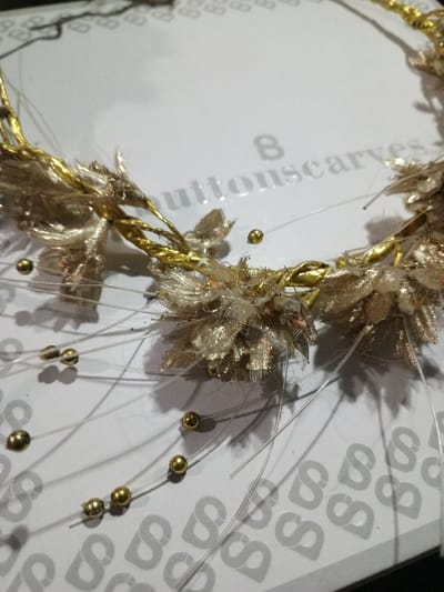 A close-up shows a golden flower crown resting on a white surface. The crown features fabric flower accents and small golden beads attached to thin, transparent strands. The surface has a subtle grey pattern of repeated "S" shapes and the text "buttonscarves" is faintly visible, suggesting the crown is displayed on its packaging.