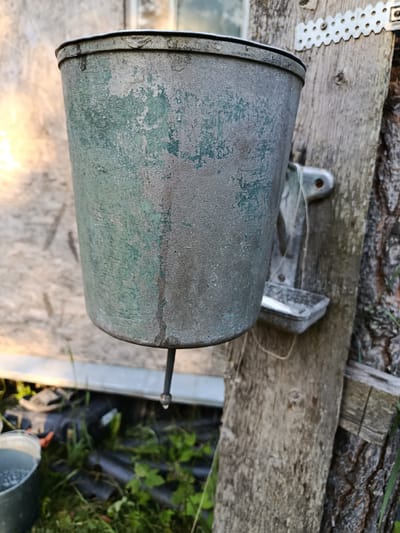 An old, weathered green metal bucket is mounted on a weathered wooden post, suggesting an outdoor location. The bucket has a spout at the bottom from which a single drop of water is falling. Below the spout, a small metal basin is attached to the post. The background consists of a textured wall, some greenery, and what appears to be a bucket or container on the ground. The overall impression is of a rustic, functional setup for washing or dispensing water. The image does not provide any information about the city of Apraksin, Russia, or any individuals present.