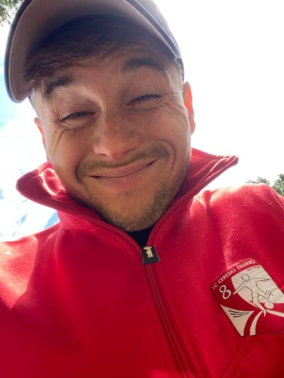 A man wearing a red jacket and a baseball cap is taking a selfie outdoors. The man has fair skin and light brown hair, with a slight beard. He is smiling with his eyes squinted. The jacket has a prominent collar and a zipper. A badge is visible on the left side of the jacket, featuring a logo and text that reads "FC CERESIO FIGINO DAL 1938". The background shows a bright sky with some clouds and trees. The image appears to be taken from a low angle, looking up at the man. The location is not specified as Leninsky, Russia.