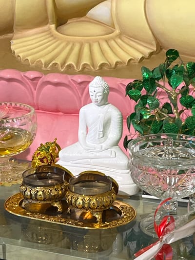 The image shows a shrine or altar, likely within a Buddhist temple or home in Katunayake, Sri Lanka. A white Buddha statue, seated in meditation, is the central figure. To its left, a glass bowl contains a yellow liquid, possibly water or oil. In front of the Buddha statue, a golden tray holds two ornate bowls, each containing a dark liquid. To the right of the Buddha, a decorative tree with green leaves is visible, and a crystal bowl filled with water sits behind it. The backdrop features elements of a larger Buddha statue, with yellow and pink decorative folds. The overall scene suggests a place of worship or devotion, adorned with offerings.