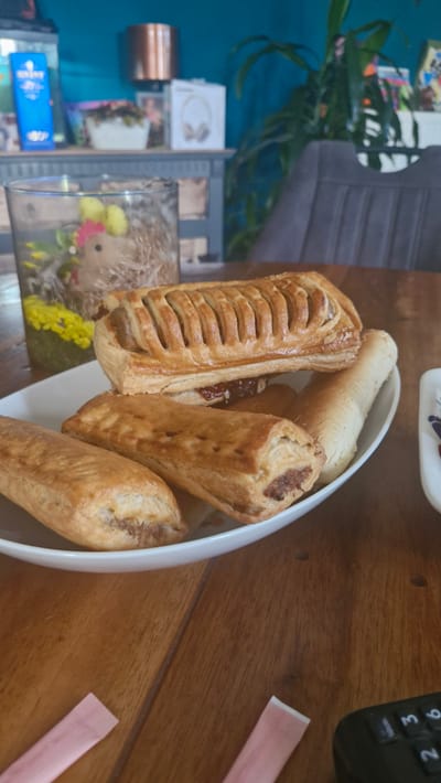 A close-up, slightly angled shot shows a white oval dish filled with several golden-brown pastries on a wooden table. The pastries appear to be sausage rolls, with some showing a flaky, layered pastry and others a smoother, baked dough. One pastry is sliced open, revealing the meat filling. A light-colored bun or breadstick is also visible in the dish. In the background, to the left, a blurry view shows a teal-colored wall and shelves with various items, including what looks like a small decorative hedgehog with yellow flowers. To the right, a portion of a black remote control with buttons labeled "9", "6", "3", and "2" is visible. Two pink paper wrappers, possibly for sugar or cutlery, lie on the table near the remote. The lighting suggests it could be daytime, possibly indoors in a cafe or home setting. No people are visible.