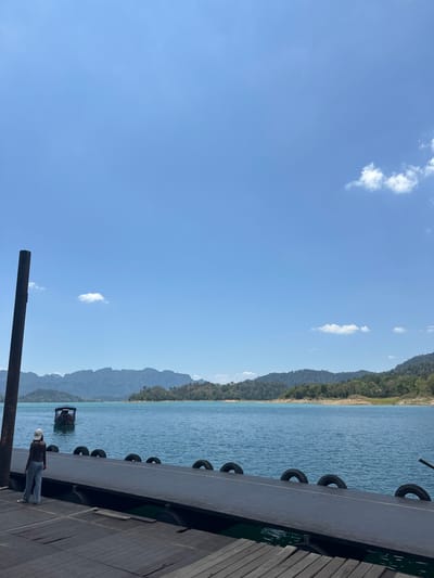 The image depicts a daytime scene at Cheow Lan Lake in Ban Ta Khun, Thailand. The composition is oriented vertically, with the sky occupying a significant portion of the frame. The sky is blue with scattered white clouds. Below the sky is a view of a large body of water with forested mountains in the background. A portion of a wooden dock extends into the frame from the right, with black tires affixed to its side. A person wearing a white hard hat, dark shirt, and blue jeans stands on the dock. A small structure is visible on the water.