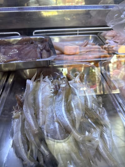 The image shows a display of fish and other seafood in what appears to be a refrigerated case, located in Villa General Belgrano, Argentina. The fish are small and silver, stacked on top of each other. To the left, in separate compartments, are other types of seafood, including what appear to be chicken and calamari. The display is likely in a market or fishmonger's shop. The fish appear to be on ice, or a similar cooling substance, to maintain freshness. The scene is viewed through a reflective surface, possibly the glass of the refrigerated case.
